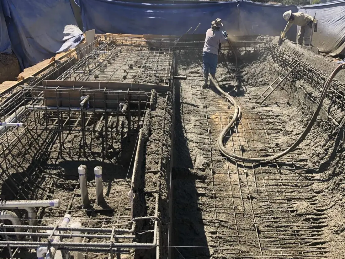 Crew spraying gunite over rebar with plumbing lines standing up in the pool floor