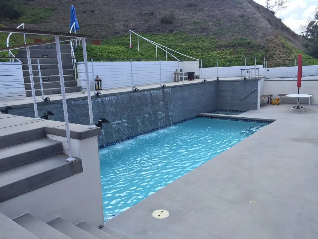 Hillside pool with a gray tile water wall and a concrete deck with steps