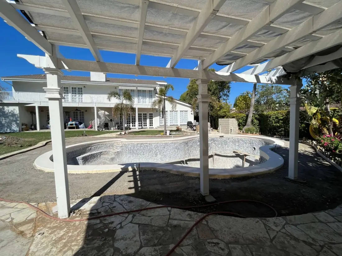 Kidney-shaped pool stripped for resurfacing under a white pergola