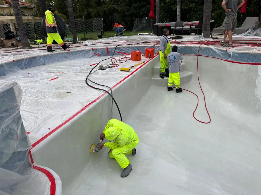 Crew in protective suits preparing a drained pool shell, with the deck masked in plastic