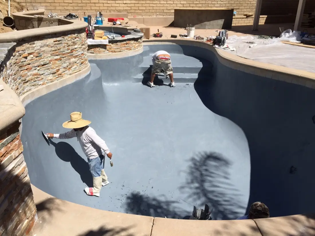 Workers coating the inside of a drained kidney-shaped pool shell during resurfacing