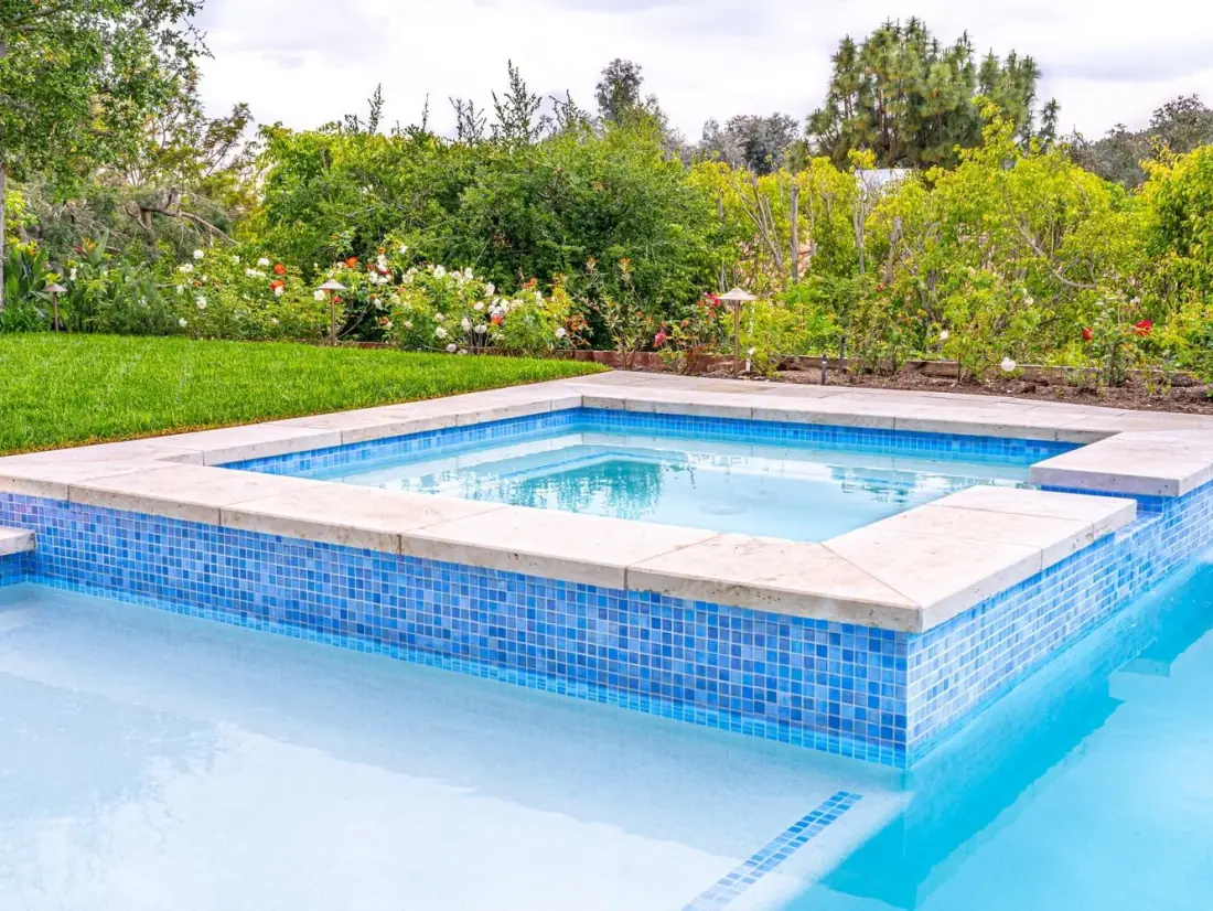 Raised spa with a blue glass mosaic tile wall and stone coping beside a pool