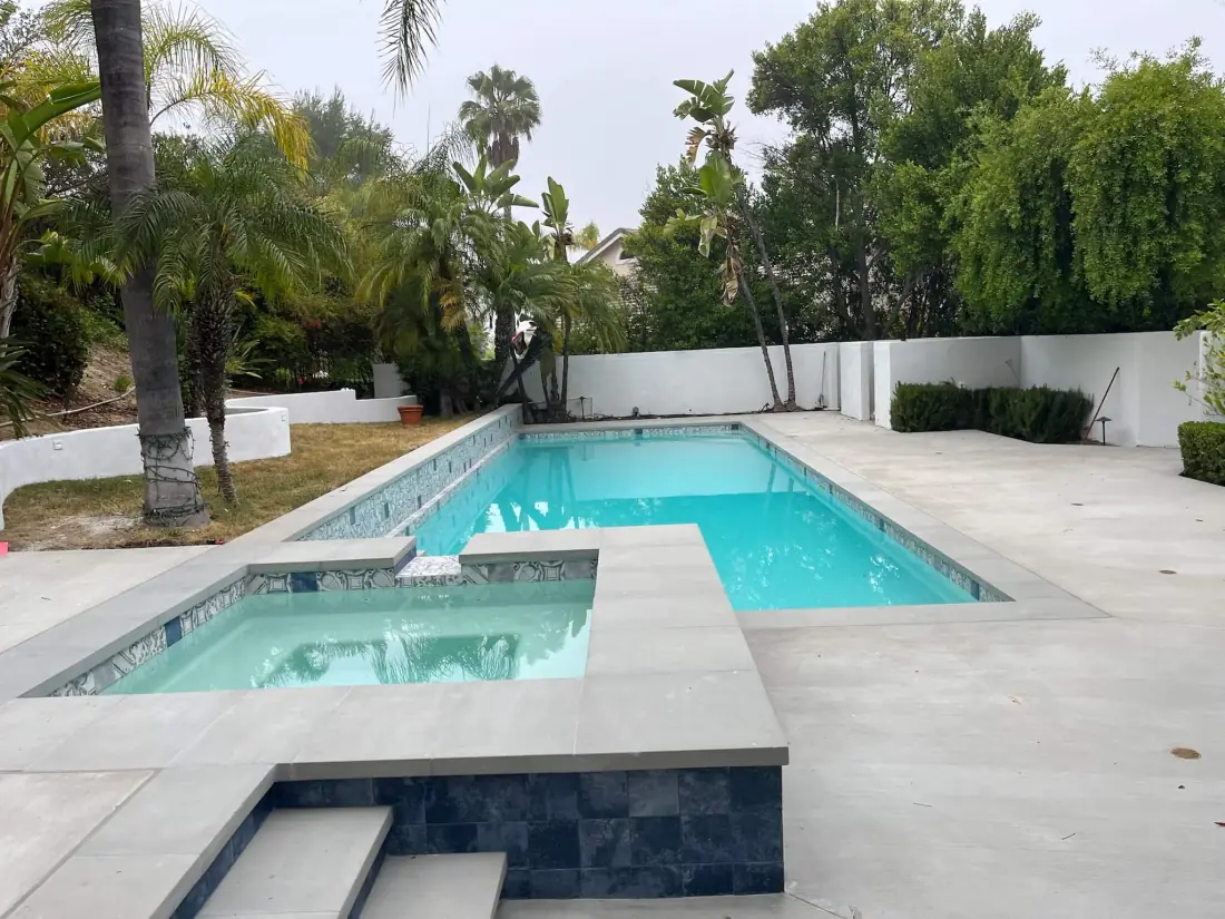 Rectangular backyard pool with an attached raised spa, dark tile band and a light stone deck